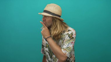 Hasır şapkalı ve çiçekli tişörtlü bir adam sessiz kalmak için dudaklarını parmaklıyor. Stüdyodaki deniz mavisi duvarına karşı..