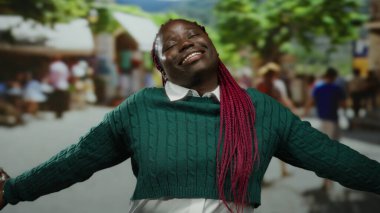 Örgü ören Afrikalı Amerikalı kadın ağaçlar ve insanlarla çevrili güneşli bir açık hava parkında kollarını açmış neşeyle gülümsüyor..