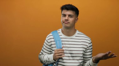 Sırt çantalı genç adam turuncu bir arka planda duruyor, düşünceli ve anlamlı görünüyor, çizgili bir gömlek giyiyor..