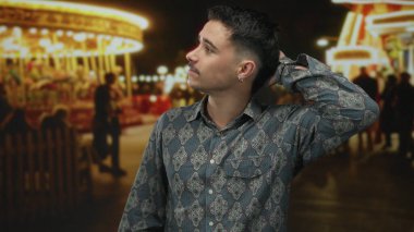 Genç İspanyol adam gece fuarında arka planda atlıkarınca ışıklarıyla eğleniyor, desenli gömlek giyiyor ve canlı park ortamında düşünceli bir şekilde poz veriyor..