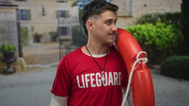 Cankurtaran tişörtlü genç İspanyol adam şehir caddesinde şamandıra ile, canlı şehir ortamında yemyeşil ile gülümsüyor.