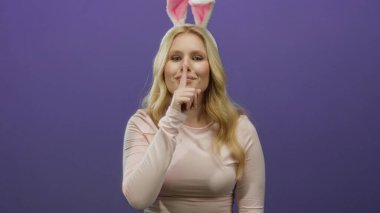 Tavşan kulaklı genç sarışın kadın canlı mor bir arka plana karşı şşşş hareketi yapıyor, neşeli ve esprili bir atmosfer yayıyor..