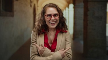 Tarihi bir üniversite koridorunda el kol hareketi yapan, şaşırdığını ve merakını dile getiren, kırmızı gözlük takan ve kapalı bir akademik ortamda bej ceket giyen bir kadın..