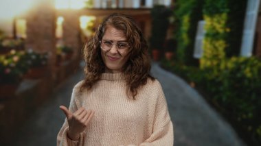 Şehir sokaklarında kadın süveter ve gözlük takıyor. Belirsizliği belirten başparmaklarıyla ifade ediyor. Çevresi bulanık yeşillik ve kentsel mimari ile çevrili..