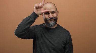 Kel kafalı, sakallı, siyah tişörtlü, komik yüzlü, parmağıyla işaret eden, kahverengi duvarlı ezik işareti yapan bir adam.