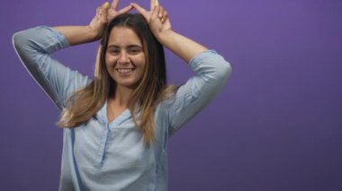 Elleri tavşan kulağı yapan kadın jest yapıyor, gözleri kapalı ve gülümsüyor, mor bir stüdyoda açık mavi gömlek giyiyor; neşeli bir neşe.