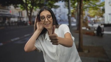 Gözlüklü ve beyaz gömlekli bir kadın. Kulaktan kulağa dinliyor ve parmaklarını kaldırıyor. Ağaç ve binanın yanında, iyimserlik merakı..