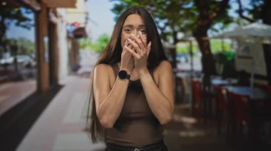 Uzun saçlı bir kadın, bir restoranın dışındaki terasta iki eliyle ağzını kapatıyor, kamera dışında bir şey izliyor; sürpriz..