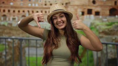Roma harabelerinde gülümseyen ve el kol hareketi yapan genç bir kadın. Açık havada, gündelik şapka ve güneşli gökyüzünün altında uzun kahverengi saçlı yeşil bir gömlek giyiyor..