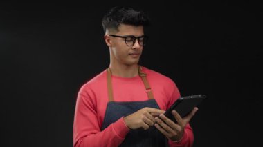 Gözlüklü ve önlüklü genç adam karanlık arka plan karşısında tabletle etkileşim içinde modern teknolojiyi ve moda tarzını sergiliyor güzellik ve basitliği vurguluyor.