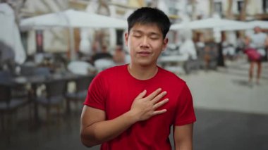 Kırmızı tişörtlü genç Çinli adam, arka planda bulanık insanlarla dolu hareketli bir kahve dükkanının terasında duygusal jestler yapıyor ve şehrin sosyal atmosferini yakalıyor.