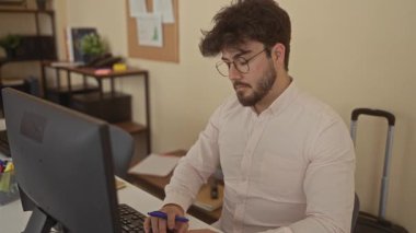 Ofis odasında bilgisayar başında çalışan sakallı ve gözlüklü genç İspanyol bir adam masa, raflar ve arka planda evrak işleriyle profesyonelce iç mekan ayarları yapıyor..