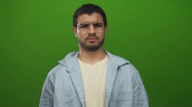 Gözlüklü İspanyol genç adam iddialı poz verir ve parlak yeşil stüdyo ortamında parmak göğse işaret eder..