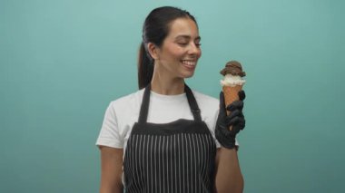 Önlüklü esmer bir kadın, eldivenli bir elinde dondurma külahını tutuyor ve turkuaz arka planlı bir stüdyoda ona gülümsüyor..