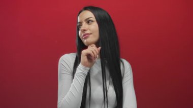 Beyaz tişörtlü genç İspanyol kadın canlı kırmızı duvara poz veriyor. Arka planı izole edilmiş. Düşünceli bir yüz ifadesi var.