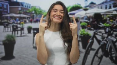 Güneşli İspanyol kadın gülümseyen ve işaret parmaklarını yukarı doğru gösteren kalabalık sokakta bulanık yayalar arasında; genç neşe.