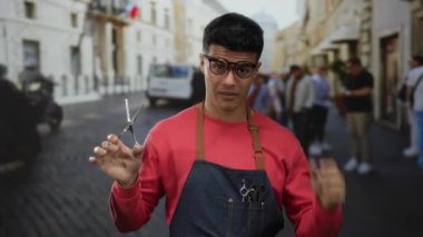 Gözlüklü ve önlüklü genç adam hareketli bir şehir caddesinde berber yeteneklerini gösterirken elinde makasla enerjik bir şehir ortamında.
