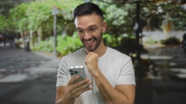 Yeşil bir parkta akıllı telefona gülümseyen, güneşli bir günde açık havanın tadını çıkaran, doğanın kucağında bir mutluluk ve rahatlama anı yakalayan genç bir adam..