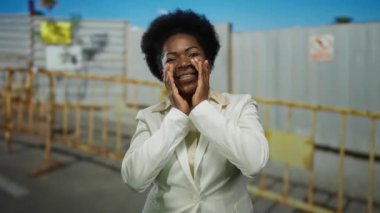 İnşaat alanında beyaz ceketli, açık mavi bir gökyüzünün altında metal çitlerle çevrili bir kadın bağırıyor.