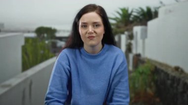 Mavi kazaklı kadın şehir sokağında, arka planda yeşillik ve açık havada moda ve kendine güvenen vücut dili ile etkileyici bir jest yapıyor..