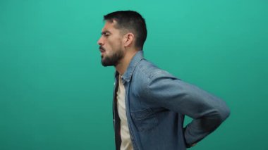 Kot ceketli genç adam yeşil bir arka planda sırt ağrısı çekiyor. Rahatsızlık ve sağlık sorunları var.