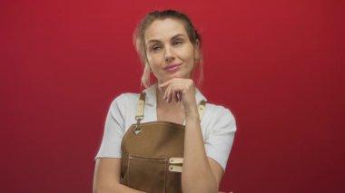 Önlüklü, kendine güvenen genç sarışın kadın koyu kırmızı bir arka plana karşı düşüncelere dalmış yaratıcı düşünce ve profesyonel zarafeti somutlaştırıyor..