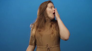 Genç beyaz kadın, mavi stüdyoda elleri ağızla kapatırken esniyor; yorgunluk uykusuzluk yorgunluğu.