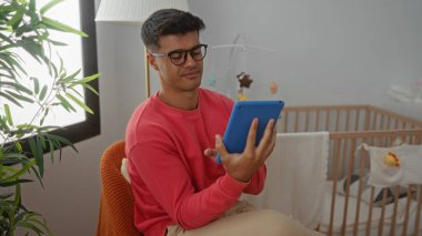 Yatak odasında tablet kullanan genç bir adam, beşiğin yanında oturuyor, modern ve rahat bir yaşam tarzını kapalı bir ortamda sergiliyor..