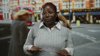 Arka planda ikonik kırmızı bir otobüsle şehir caddesinde duran gözlüklü ve gri kazaklı bir kadın..