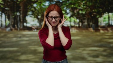 Parkta kızıl saçlı bir kadın, dışarıda gürültüden kulaklarını tıkayarak genç bir yetişkinin çevre sesine olan tepkisini çekici bir park ortamı ile doğal ortamda yakalıyor..