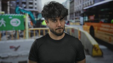 Sakallı, genç İspanyol bir adam. Yoğun bir inşaat sahasında duruyor. Çeşitli ekipman ve yapılarla..