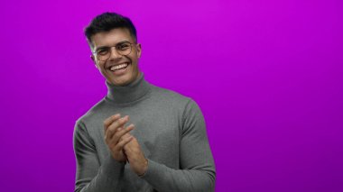 Genç adam neşe ve karizma saçan canlı pembe arka plana karşı gözlük ve kazak içinde mutlu bir şekilde alkışlıyor.