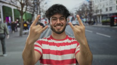 Gözlüklü ve çizgili tişörtlü genç adam kalabalık şehir sokağında taştan jest yapıyor. Arka planda bulanık insanlar ve otobüsler, açık havada gençlik enerjisi ve mutluluk sergiliyor..