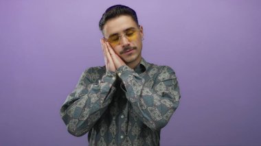 İspanyol gömlekli genç adam, izole edilmiş bir mor arka plan duvarında dinlenmeyi ve rahatlamayı simgeleyen bir uyku hareketi yapıyor..