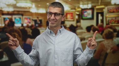 Kalabalık bir sanat galerisinde gülümseyen ve işaret eden bir adam duvarlarında canlı resimler olan ve arka planda oturan insanlar canlı bir iç mekan atmosferi sergiliyorlar..