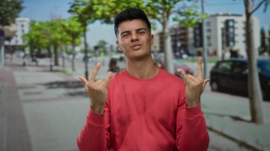 Kırmızı gömlekli genç adam şehir arka planında komik jestler yapıyor ve açık mavi gökyüzü altında modern binalar, neşeli bir güven ifadesi sergiliyor..