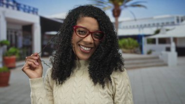 Kırmızı gözlüklü bir kadın, kıvırcık saçlarını çekerken gülümsüyor ve kendini oyuncu bir şekilde ifade ediyor..