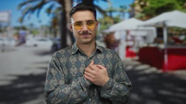 Güneşli bir şehir sokağında arka planda palmiye ağaçları ve bulanık pazar çadırlarıyla kalp hareketi yapan genç bir İspanyol..