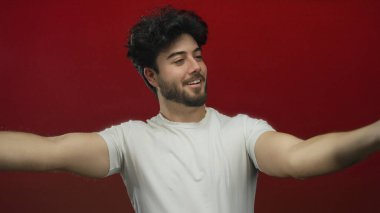 Kolları açık gülümseyen genç bir adam canlı kırmızı bir arka planda neşeli bir hoş geldin jesti sergiliyor sakallı ve günlük beyaz bir tişört giyiyor..