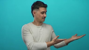 Latin kökenli genç bir adam, mavi izole edilmiş arka plana karşı elleri açık bir şey sunuyor. Günlük kıyafetler giyiyor..