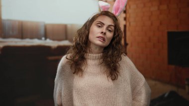 Kapalı bir inşaat sahasında tavşan kulağı takan ve arka planda tuğlalar ve borular olan bir kadın neşeli ve samimi bir ifade yayıyor..