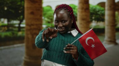 Dışarıda elinde hindi bayraklarıyla gülümseyen bir kadın, elinde kendinden emin bir jestle kültürünü sergiliyor, sonbahar kıyafetleri mimari ortamların arasında görünür..