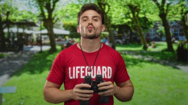 Kırmızı cankurtaran tişörtlü bir adam parkta dürbün tutuyor kameraya doğru büzüşmüş dudaklar;.