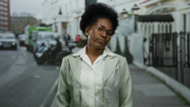 Gözlüklü bir kadın şehir caddesinde kendine güvenerek duruyor, takım elbise giyiyor, profesyonelliği ve şehir yaşam tarzını somutlaştırıyor..