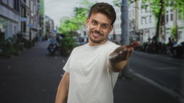 Genç adam elini davetkar bir şekilde uzatır ve kalabalık bir şehir caddesinde mobiletler ve binalarla dolu bir şekilde gülümseyerek dostça selamlar..