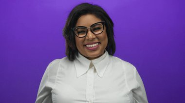 Gözlüklü genç Latin kadın sıcak bir şekilde gülümsüyor, canlı mor bir arka plana karşı jest yapıyor, şık beyaz bir gömlek giyiyor, pozitiflik ve kendine güven yayıyor..