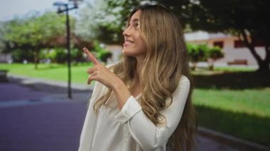 Beyaz bluzlu sarışın kadın şehir parkındaki sokak lambasını işaret ediyor..