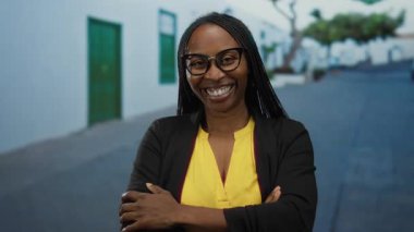 Şehir caddesinde kolları çaprazlanmış, gözlüklü ve sarı gömlekli bir kadın rahat bir şehir ortamında kendine güven ve pozitiflik sergiliyor..