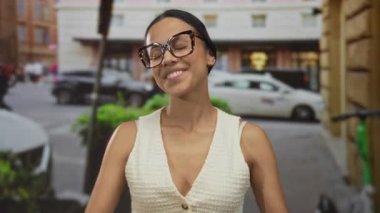 Gözlüklü, beyaz tişörtlü, kalabalık bir sokakta şükranlarını sunan, şehir ortamında mutluluk ve pozitiflik taşıyan bir kadın..