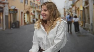 Kadın ellerini ovalıyor ve beyaz gömlek ve kemer ile dar bir kaldırım üzerinde gülümsüyor, sıcak güneş ışığı ve binalar; beklenti anı.